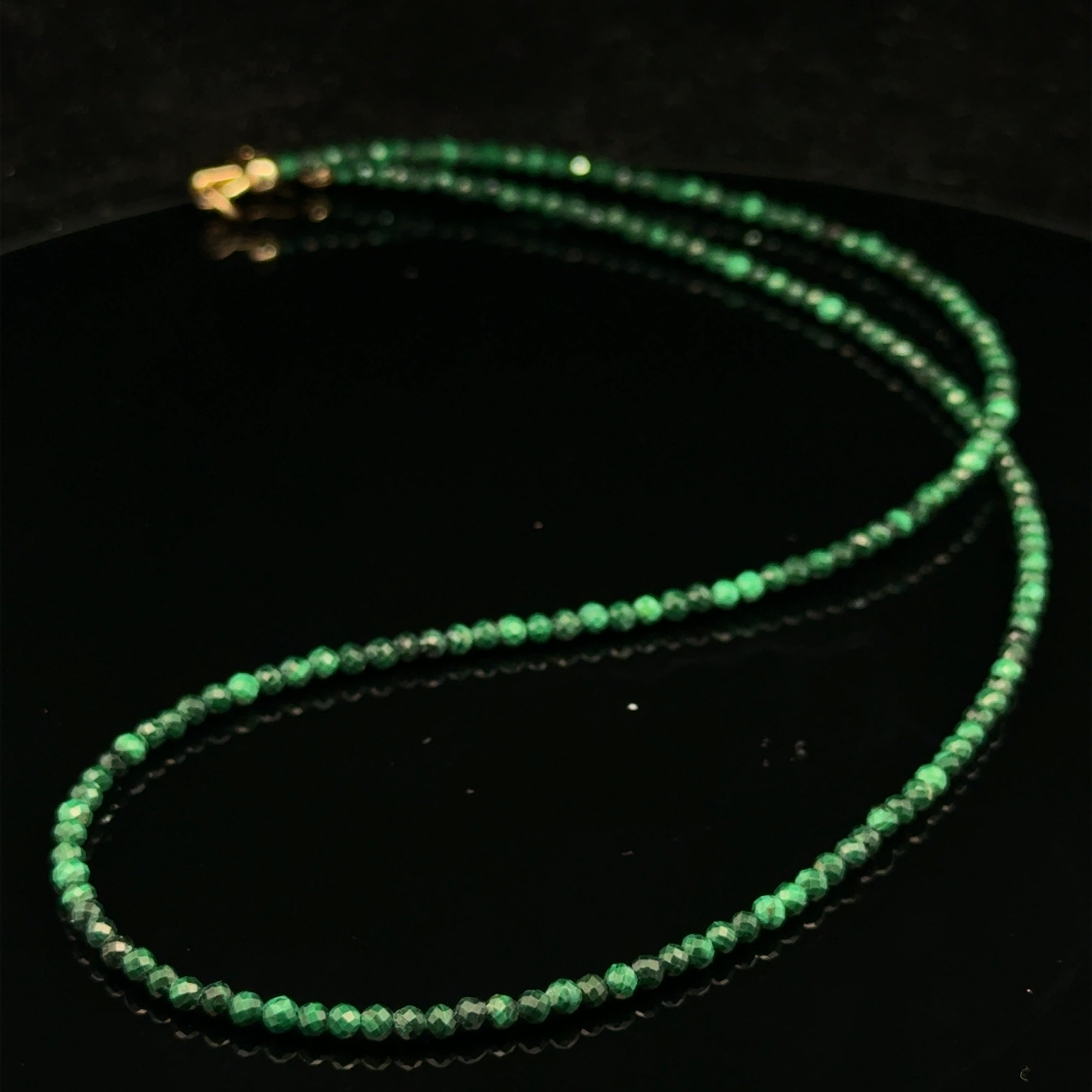 A 2mm faceted round malachite gemstone necklace with gold-filled lobster claw clasp on black background, long overview. 