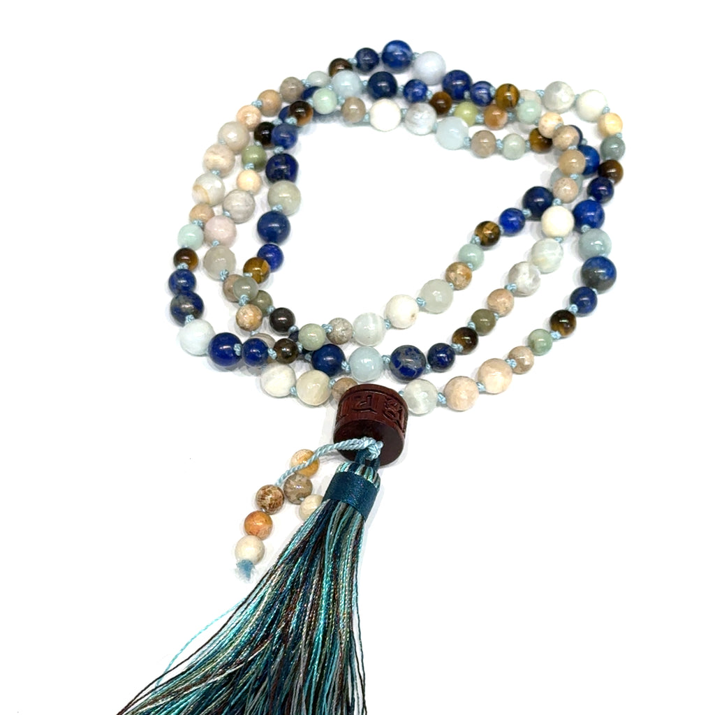 A beaded gemstone Mala necklace with a variety of colors, including blue, white, and brown, featuring a tassel end, on a white background, center, circle, close-up overview. 