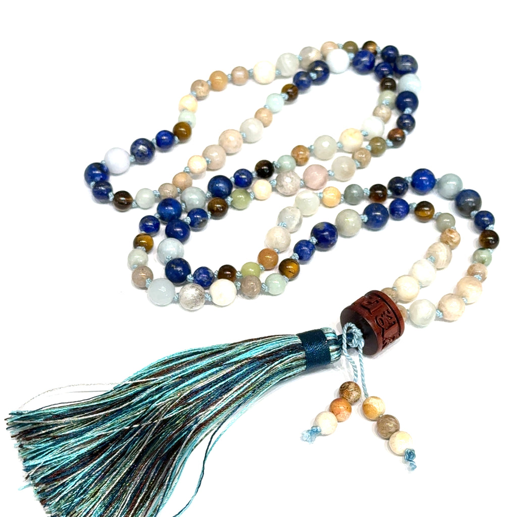 A beaded gemstone Mala necklace with a variety of colors, including blue, white, and brown, featuring a tassel end, on a white background, center front, close-up overview. 