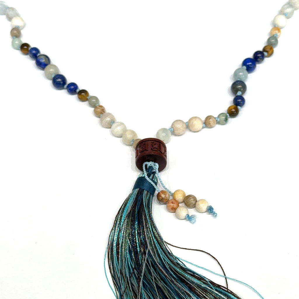 A beaded gemstone Mala necklace with a variety of colors, including blue, white, and brown, featuring a tassel end, on a white background, center, hanging, close-up overview. 