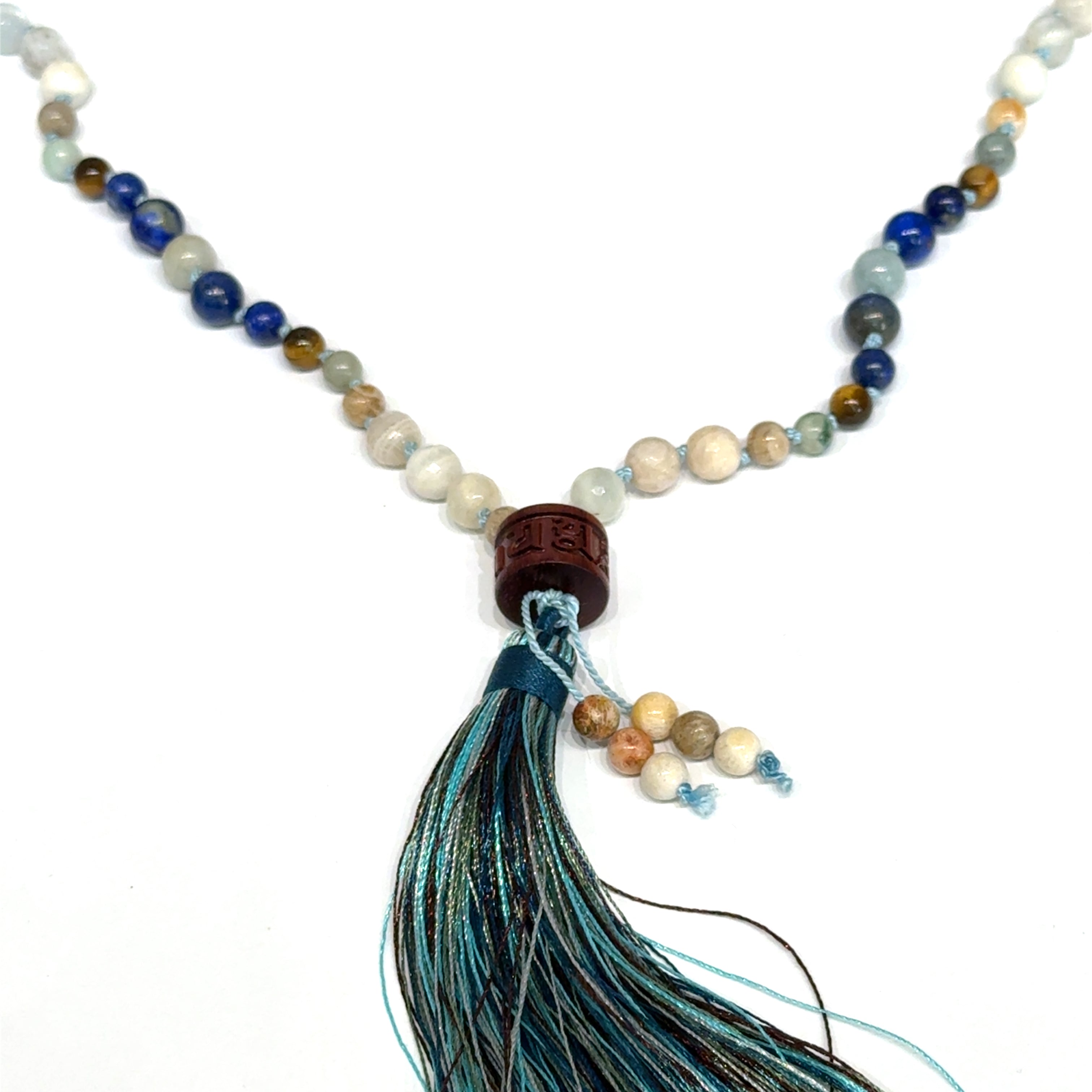 A beaded gemstone Mala necklace with a variety of colors, including blue, white, and brown, featuring a tassel end, on a white background, center, hanging, close-up overview. 