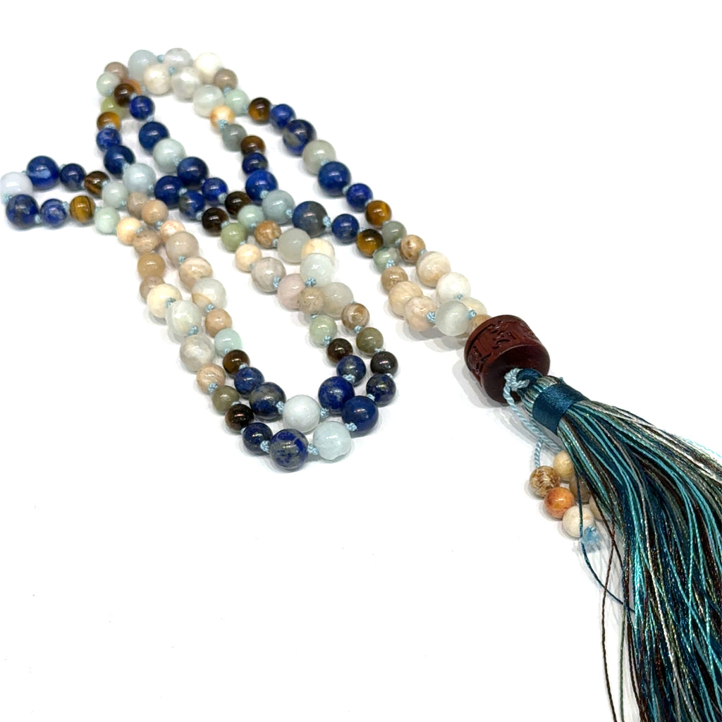 A beaded gemstone Mala necklace with a variety of colors, including blue, white, and brown, featuring a tassel end, on a white background, center, long, close-up overview. 
