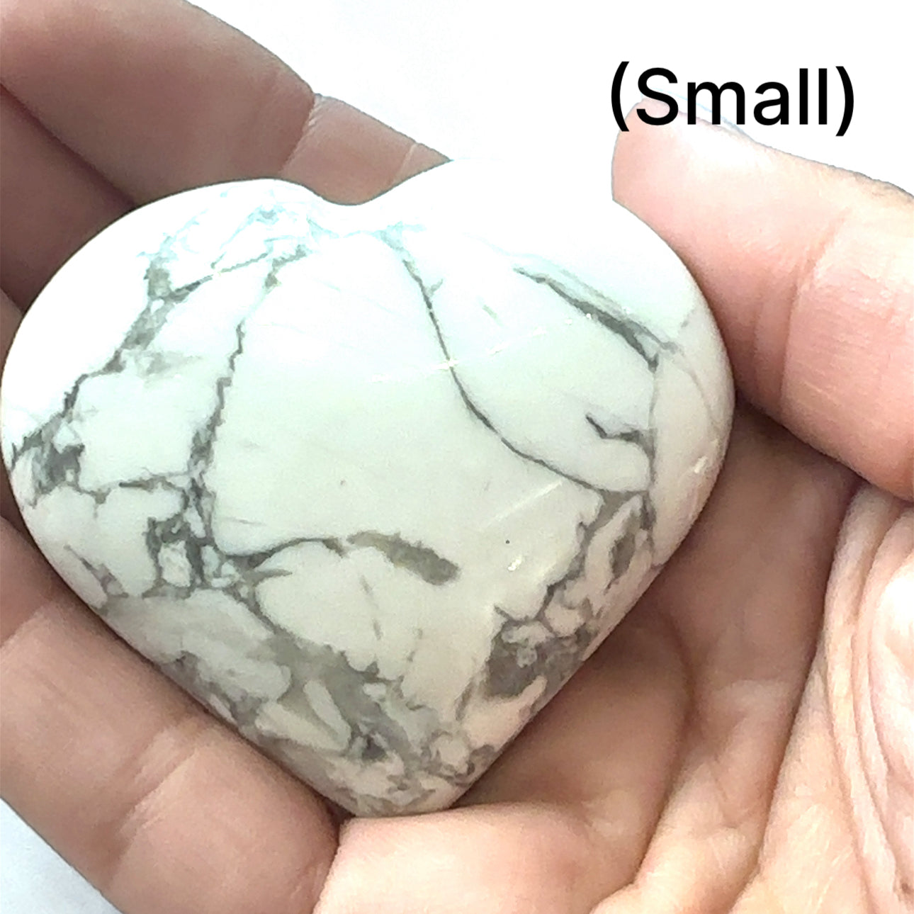 A hand holding a heart-shaped Howlite gemstone with a white and gray marbled pattern, size small, on a white background, front view.