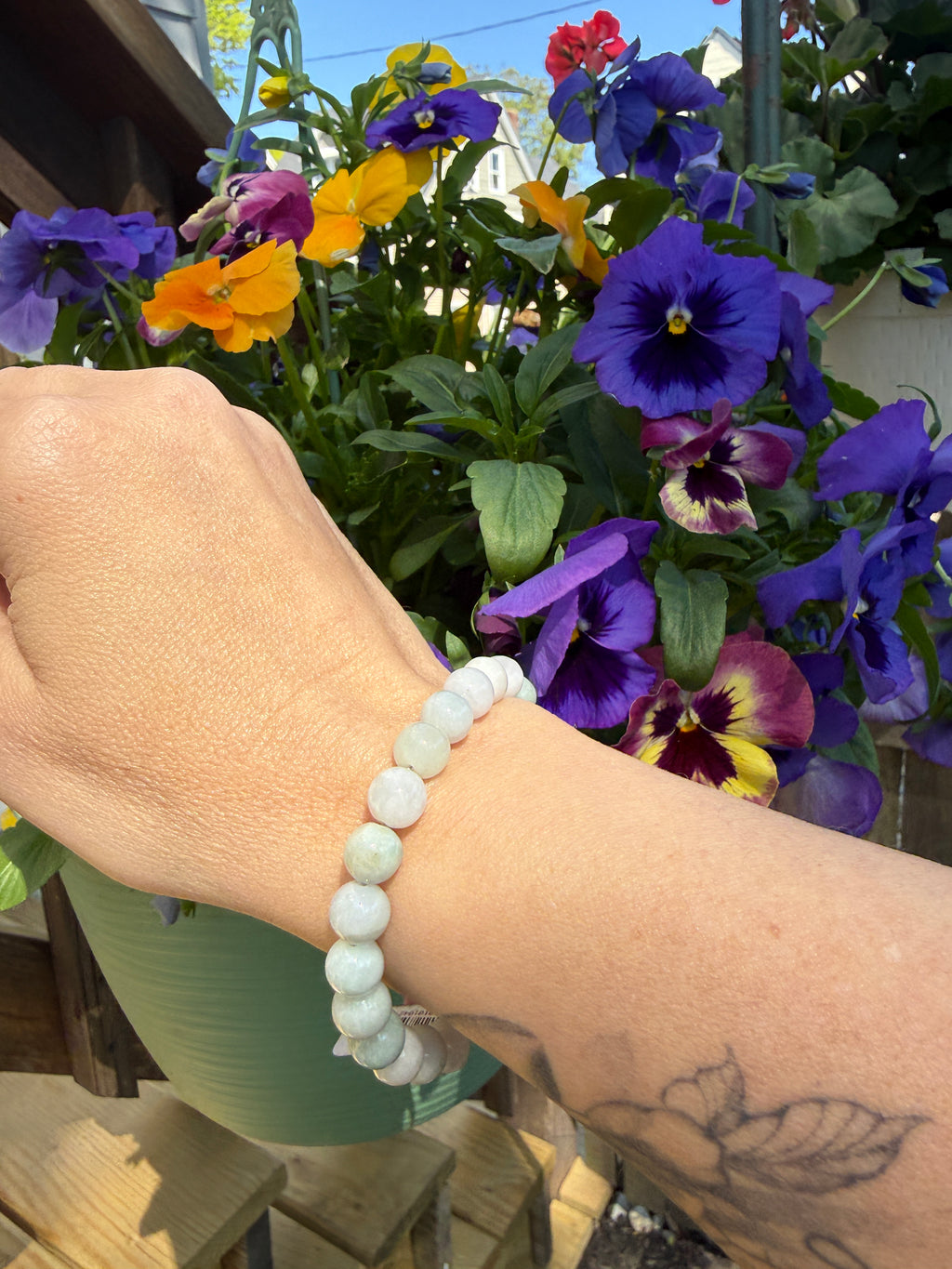 A hand wearing a Burma Jade beaded bracelet with a colorful flower arrangement in the background, front view.