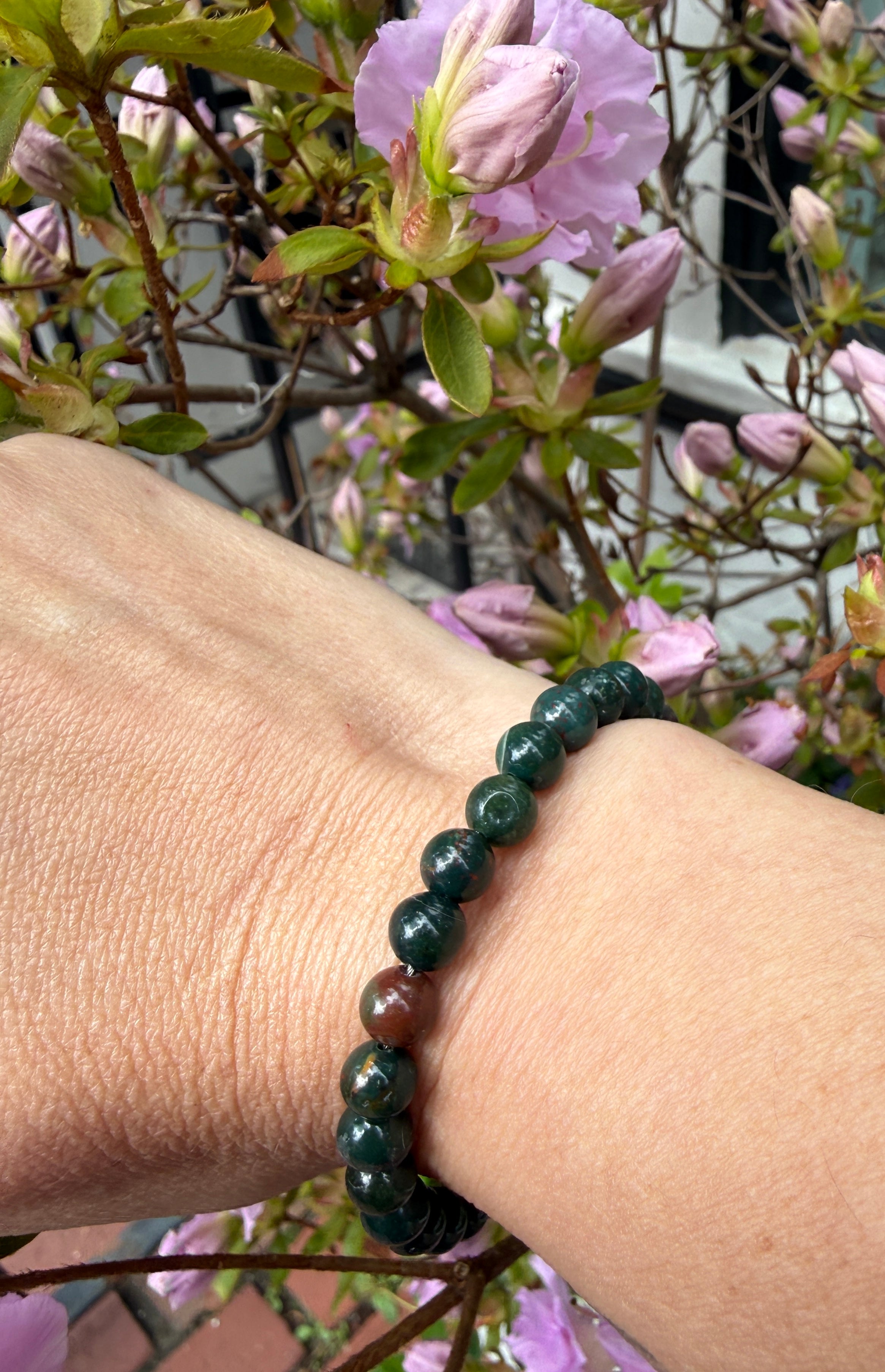 A hand wearing one 6mm Bloodstone beaded gemstone bracelet, with flowers in the background, in a front view. 