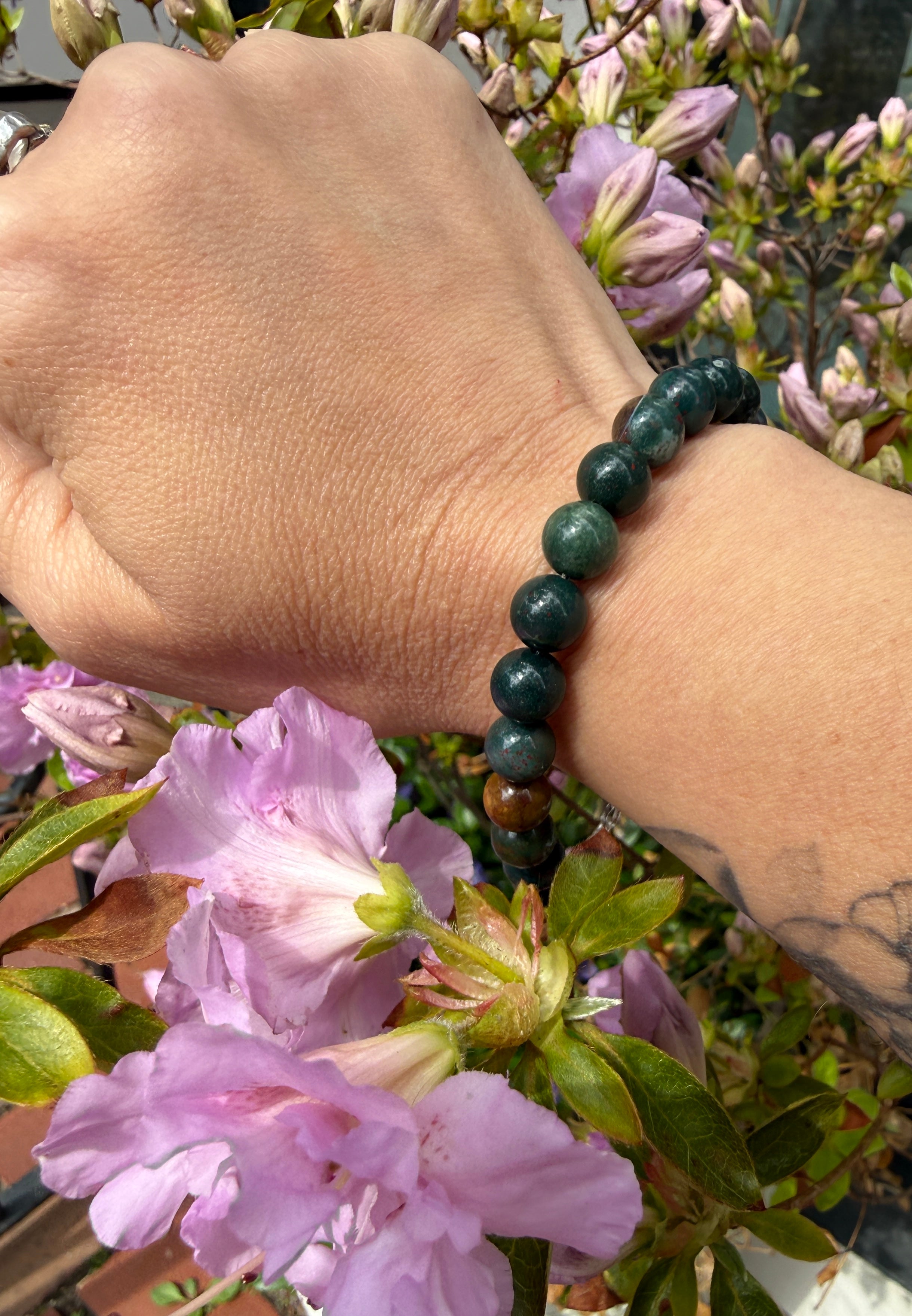 A hand wearing one 8.5mm Bloodstone beaded gemstone bracelet, with flowers in the background, in a front view. 