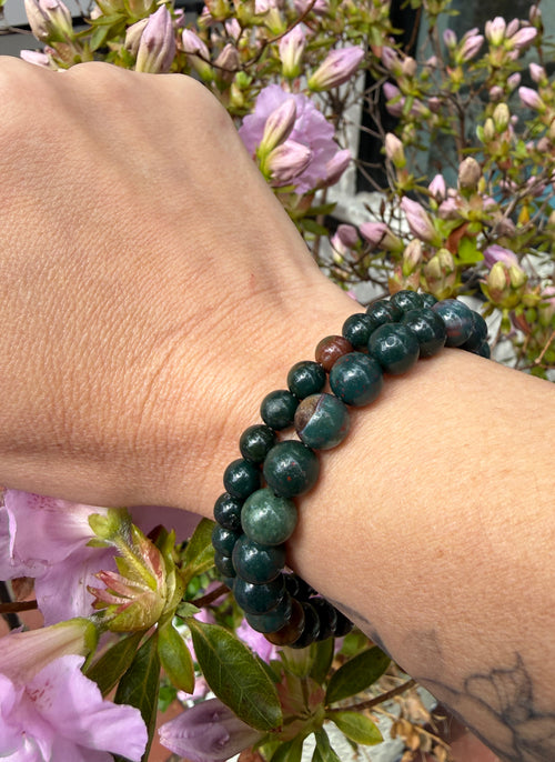 A hand wearing two Bloodstone beaded gemstone bracelets, with flowers in the background, in a front view. 
