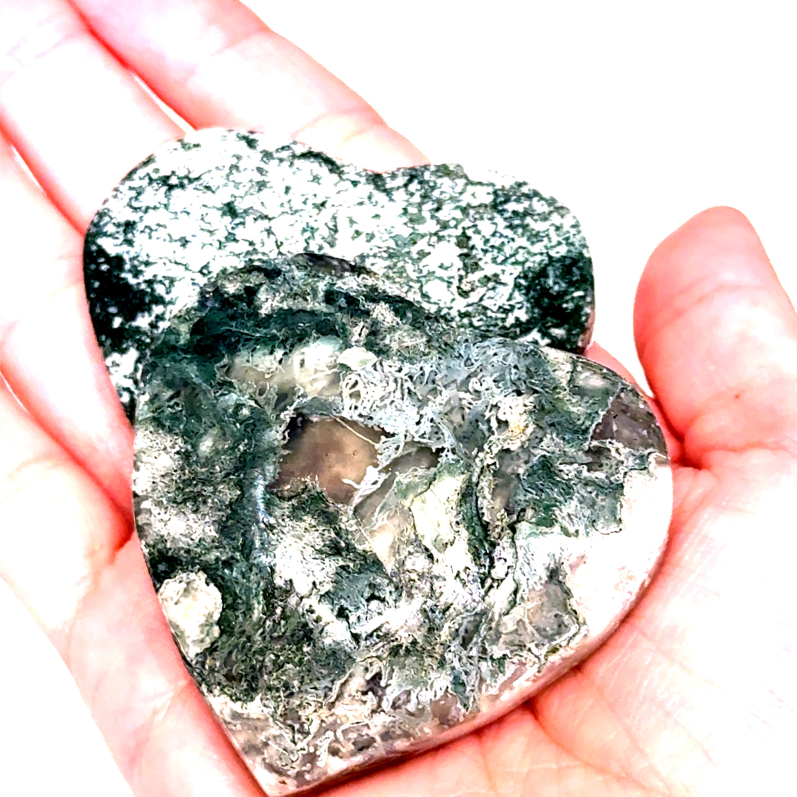 A heart-shaped Moss Agate gemstone carving held in a model's hand, displaying green and white patterns. On a white background, a close-up view.
