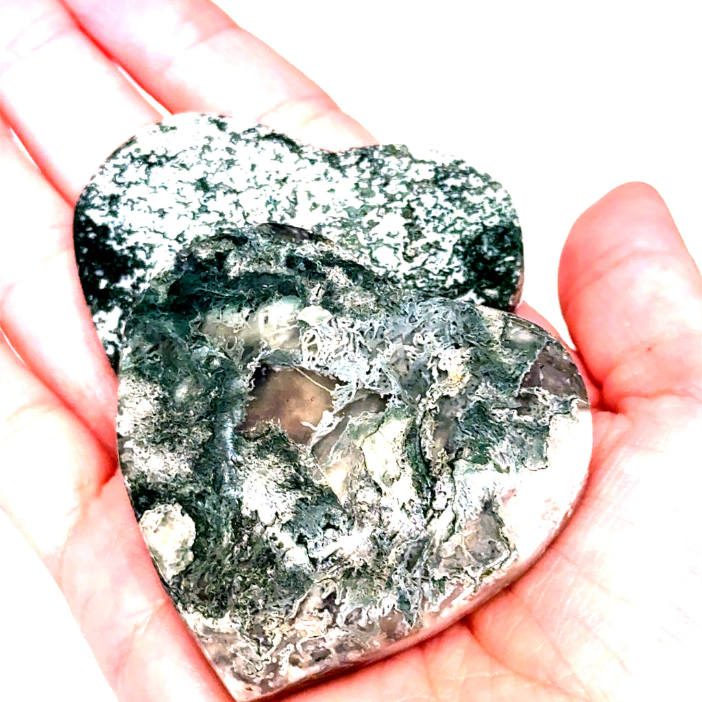 A heart-shaped Moss Agate gemstone carving held in a model's hand, displaying green and white patterns. On a white background, a close-up view.