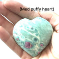 A med puffy heart-shaped Ruby Fuchsite gemstone carvings displayed in a models hand on a white background front overview.