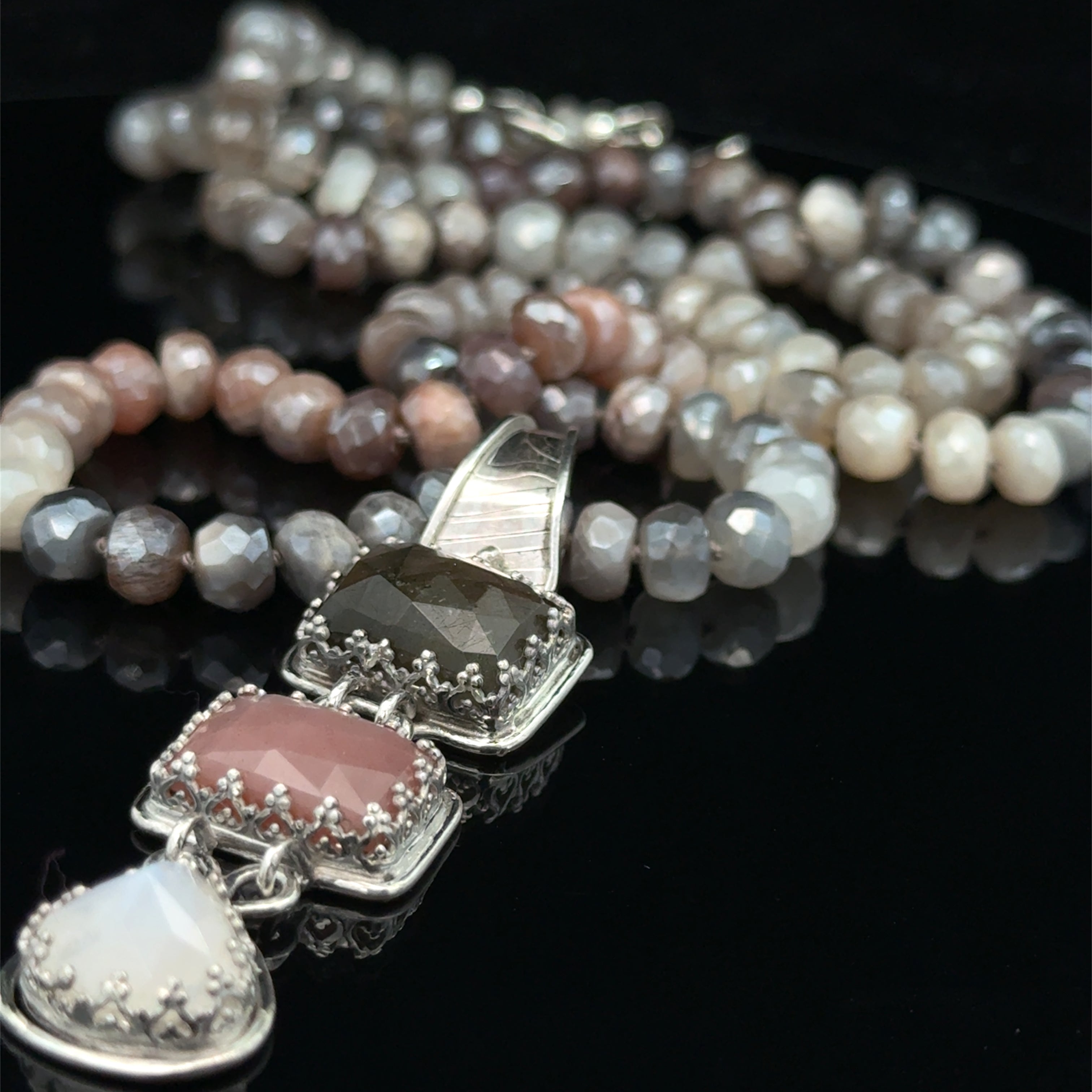 A mixed moonstone faceted roundel knotted necklace with a custom forged sterling silver with three gemstones pendant. on a black background, front overview. 