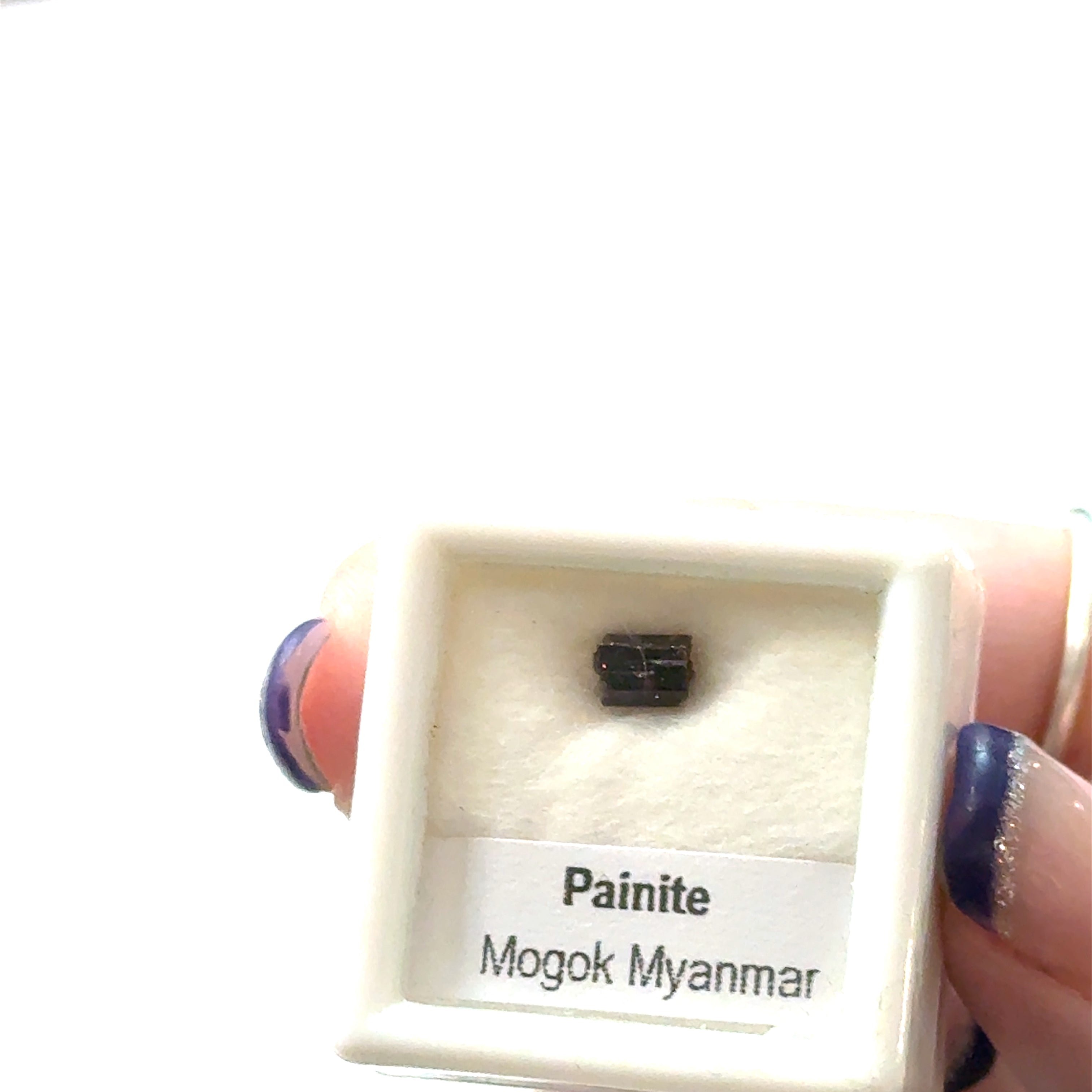 A small gemstone in a display case with 'Painite Mogok Myanmar' label and weighing  .75ct  on a white background front view. 