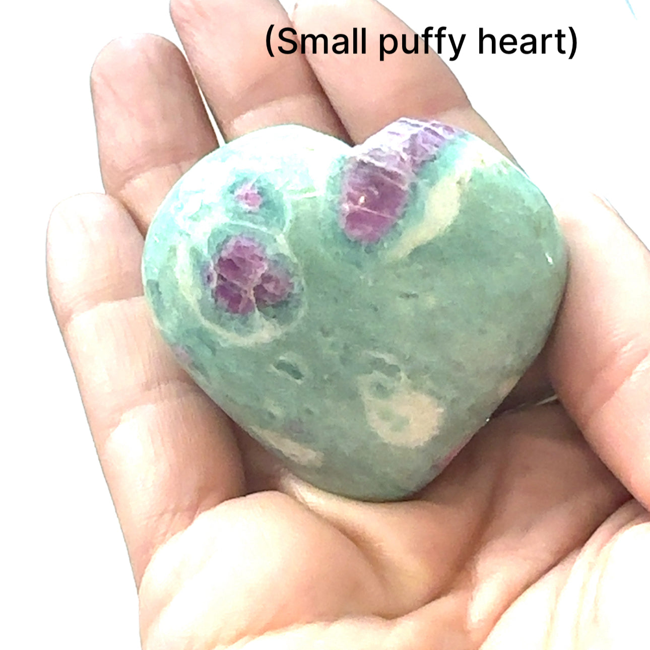 A small puffy heart-shaped Ruby Fuchsite gemstone carvings displayed in a models hand on a white background front overview.