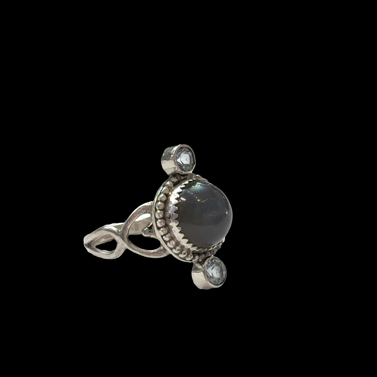 Close-up of grey moonstone adularescence glow with faceted blue topaz accents, sterling silver decorative bezel, spiritual ring on a black background, left side view. 