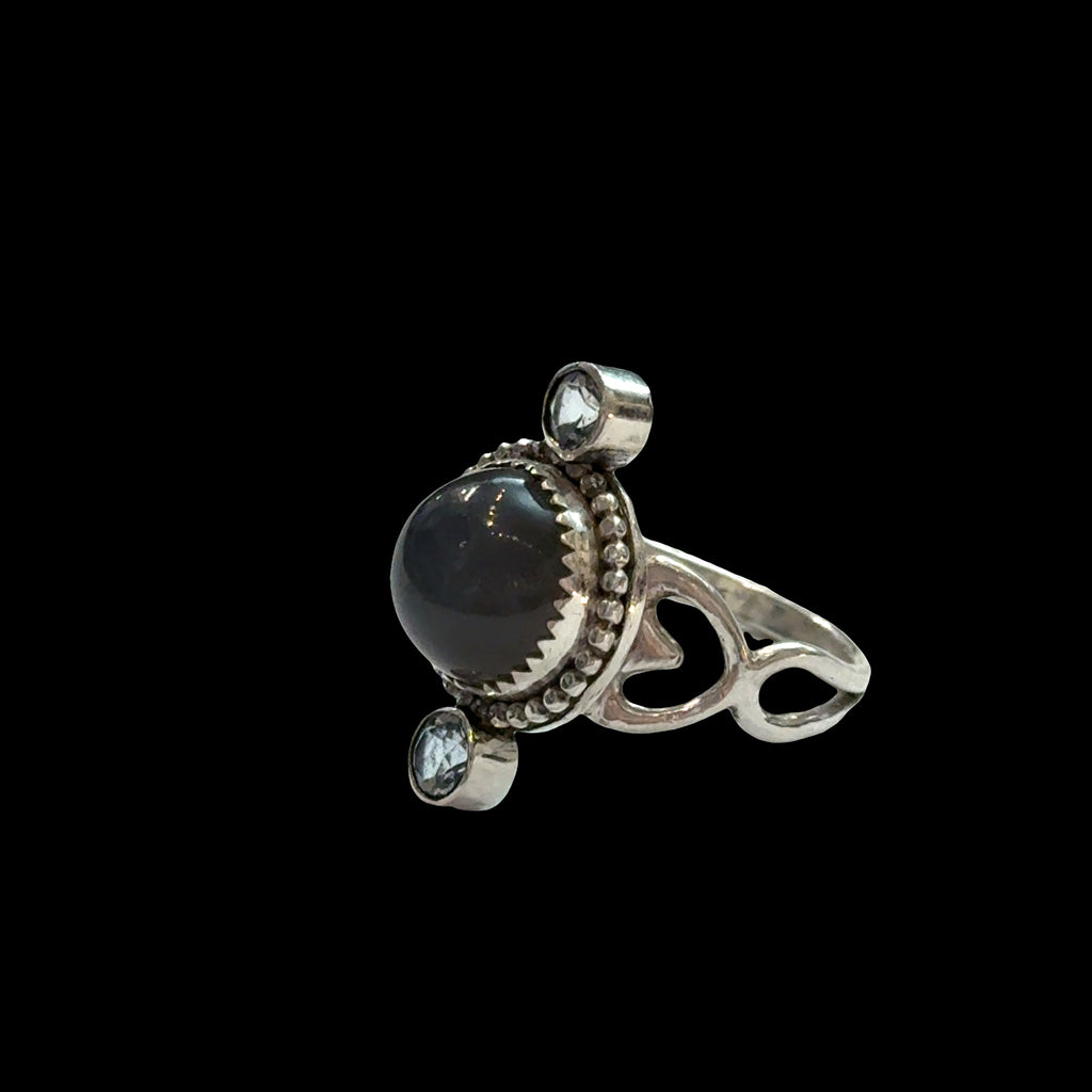 Close-up of grey moonstone adularescence glow with faceted blue topaz accents, sterling silver decorative bezel, spiritual ring on a black background, right side view.