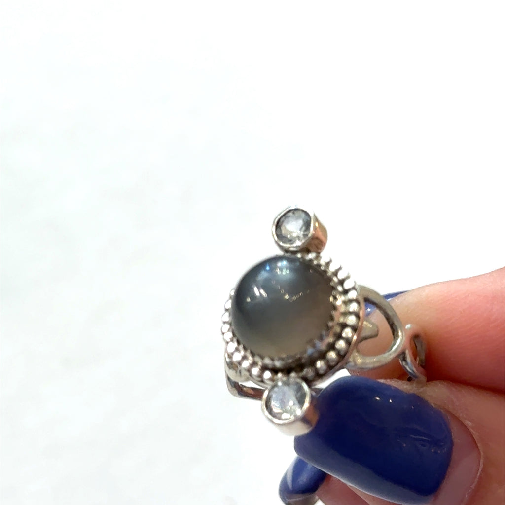 Grey moonstone and blue topaz sterling silver ring, 10mm cabochon center stone, heart-shaped bezel setting, size 6.25, intuition jewelry, on a white background, in a models finger tips front view. 