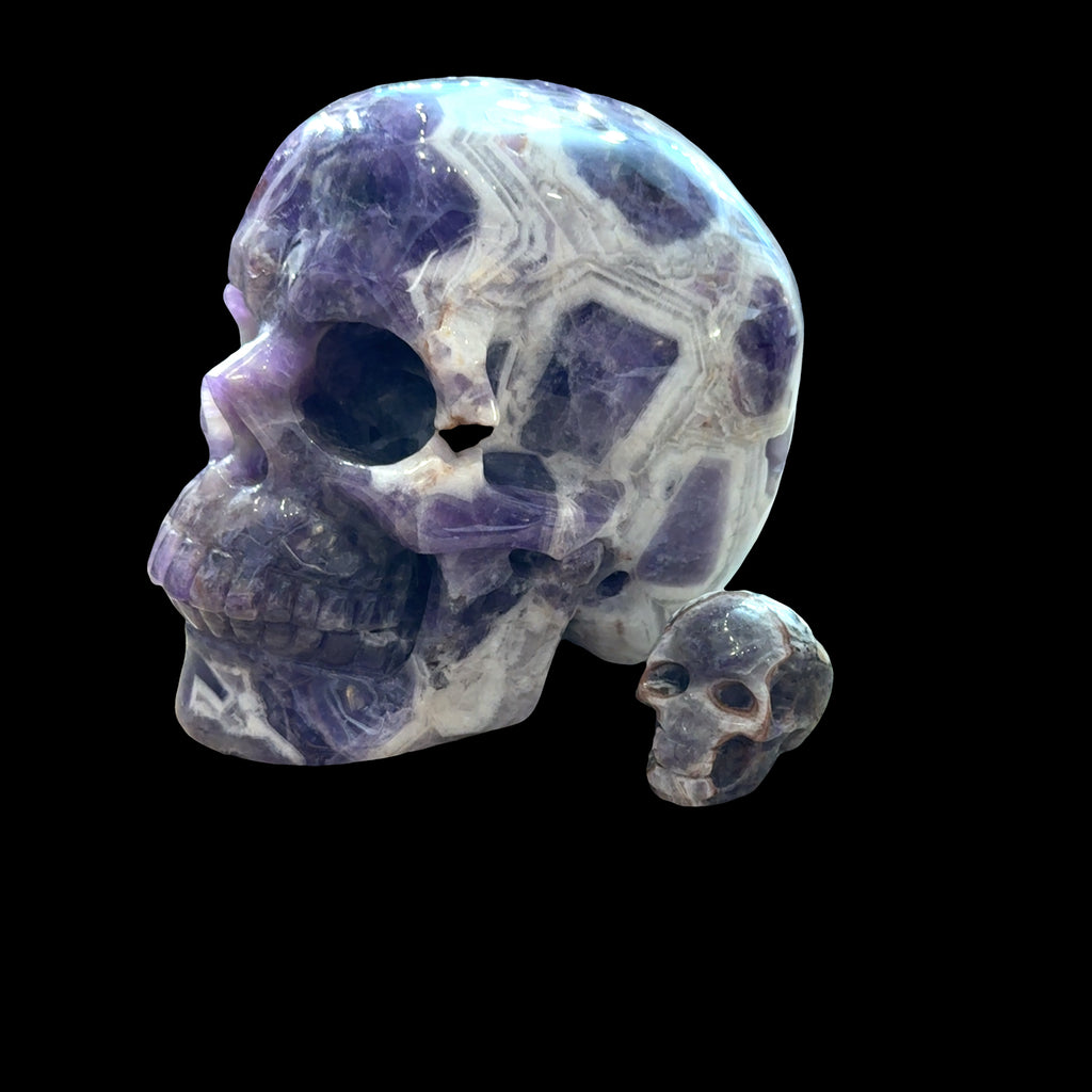 Two Trapiche Amethyst Gemstone carved skulls on a black background, left angled, front view. 