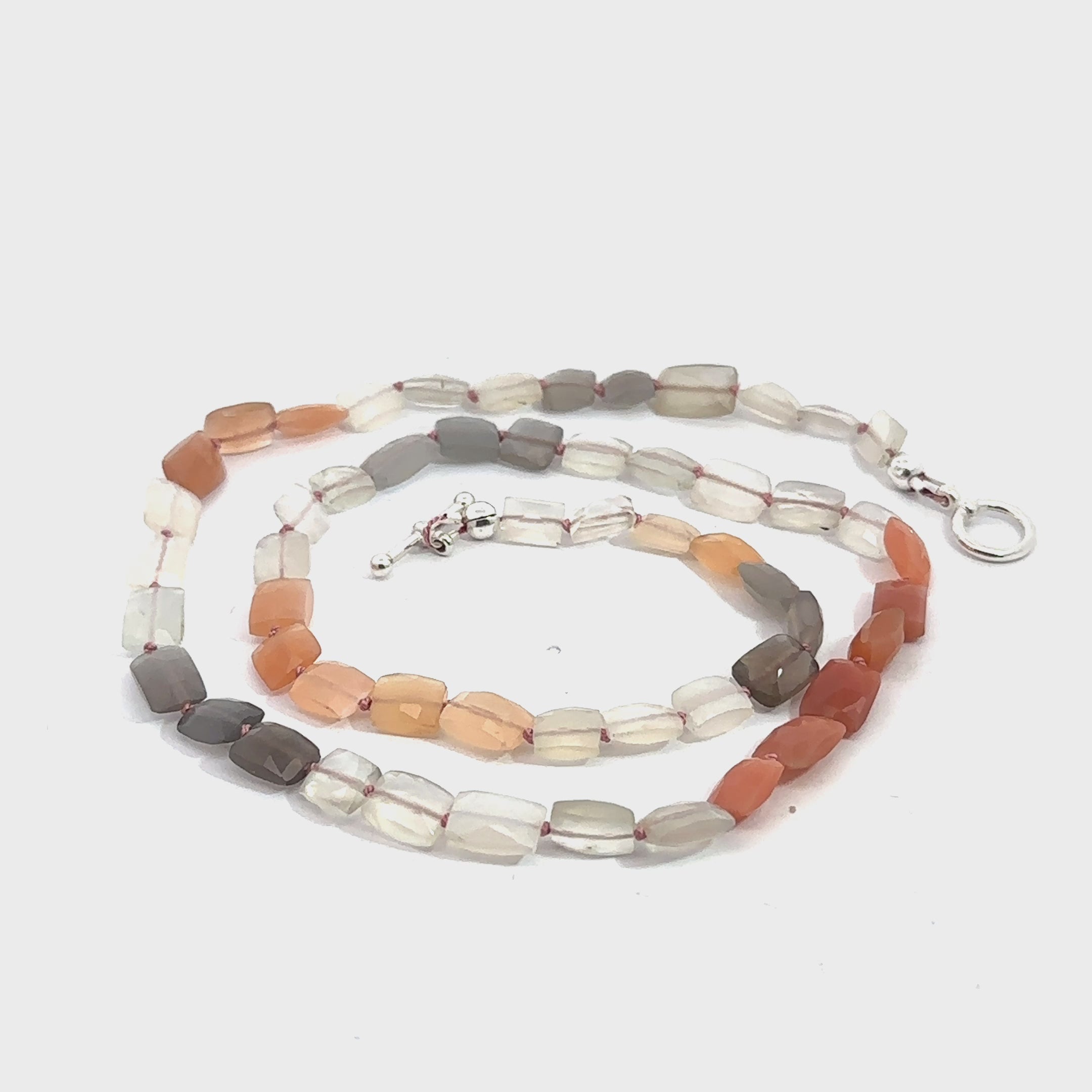 A knotted necklace made of mixed moonstone gemstones with a sterling silver toggle clasp on a white background, spiral overview. video