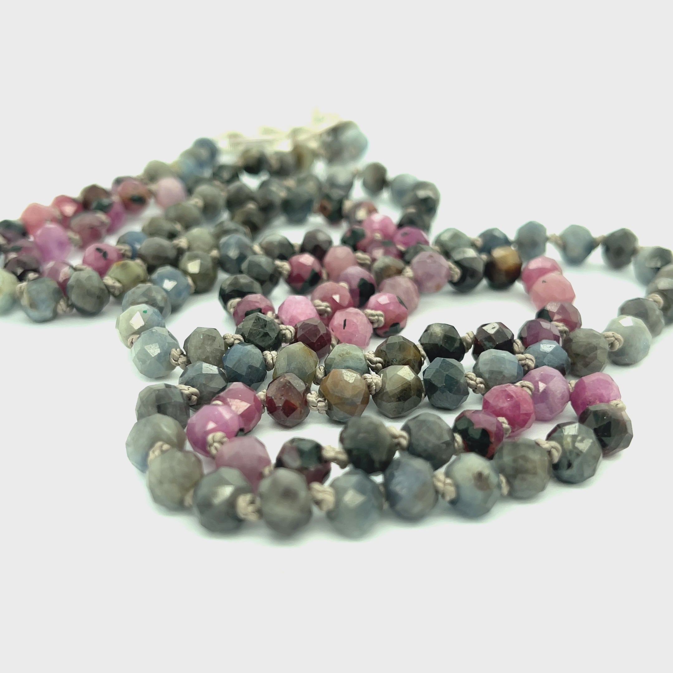 A knotted necklace featuring a heart lobster clasp, composed of faceted roundel rubies and sapphire gemstones on a white background, long center view. video