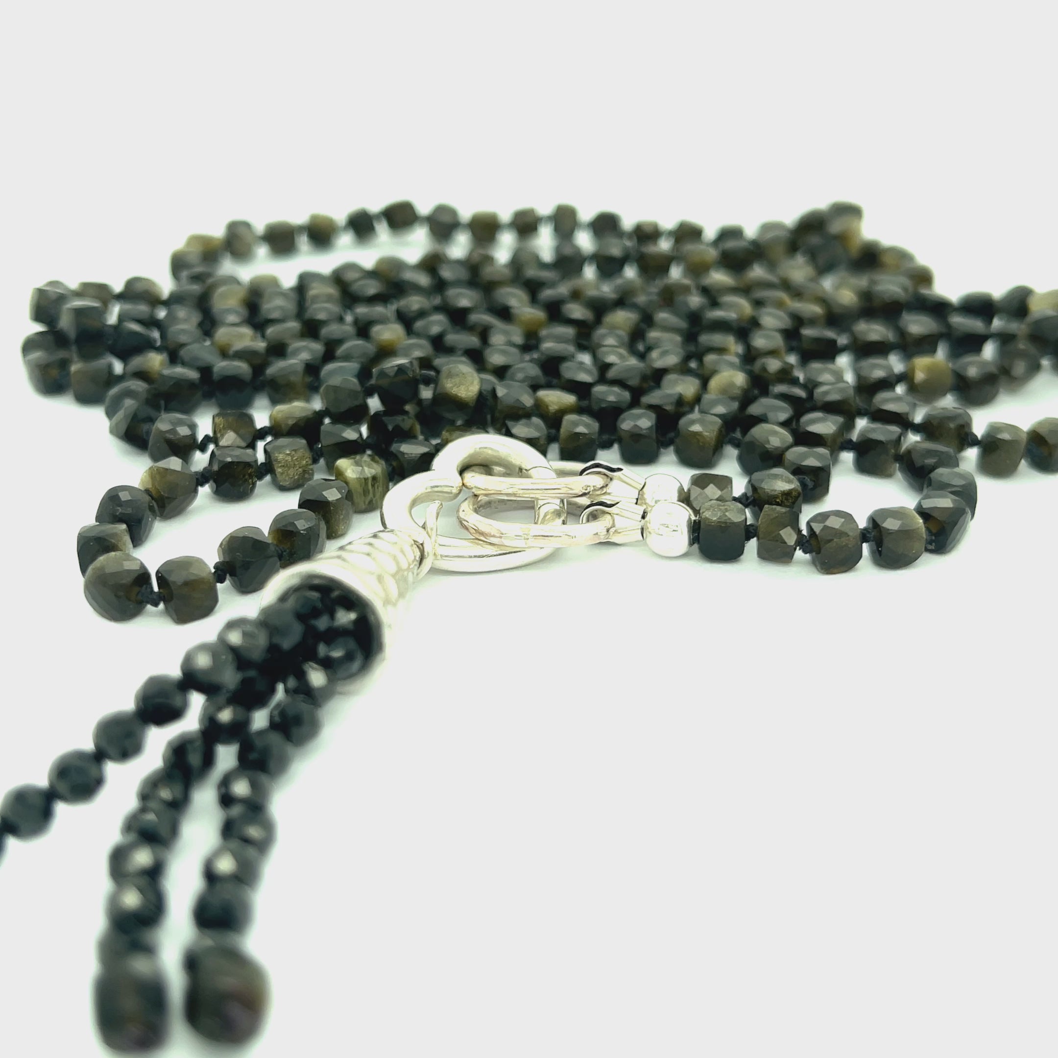 A knotted necklace made of Golden Sheen Obsidian gemstones with a tassel, featuring silver accents. On a white background, a long, center view overview.  video