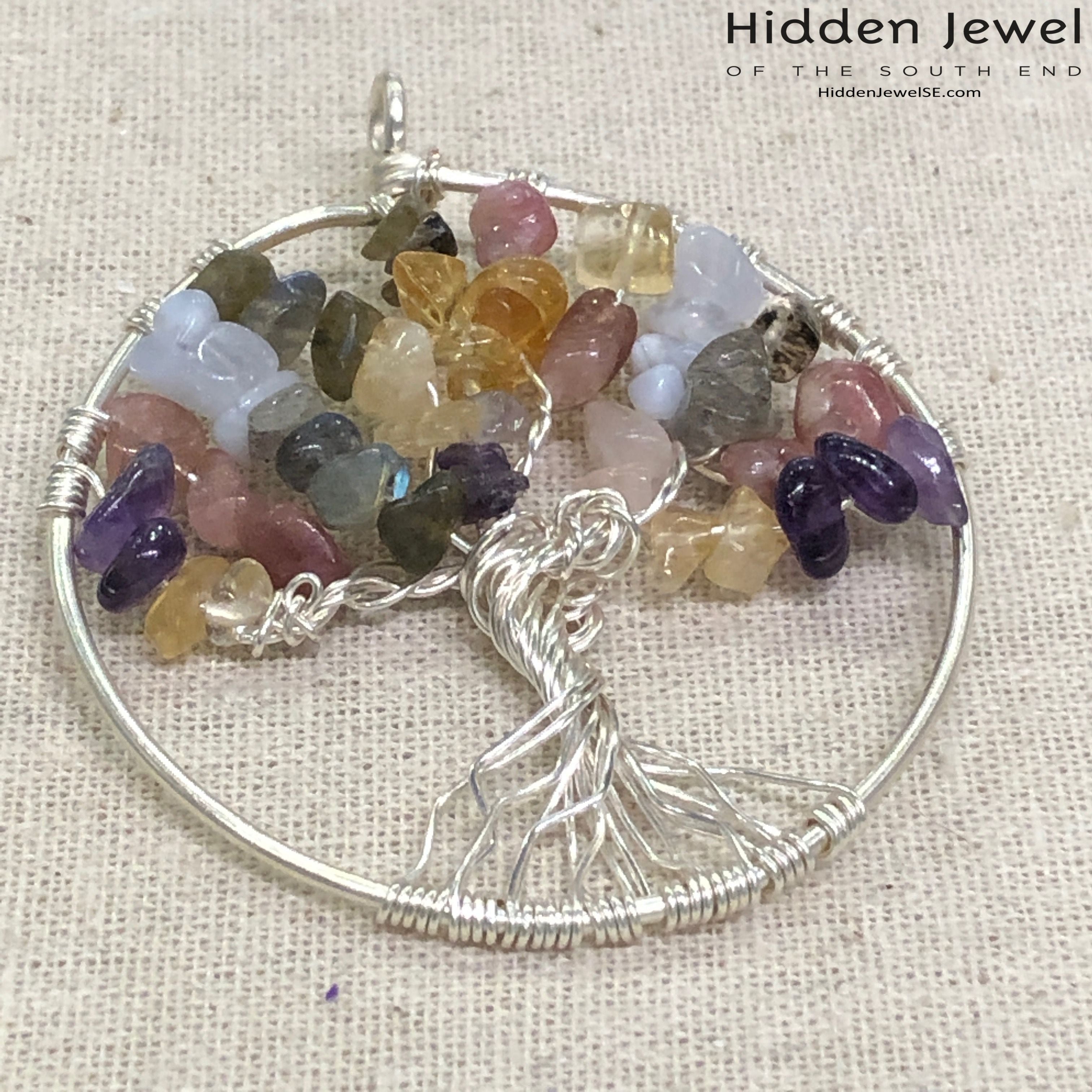 A Tree of Life pendant featuring a  sterling silver wire frame with an array of multicolored gemstones arranged in a circular pattern to form the design of a tree. on a tan background, front view.