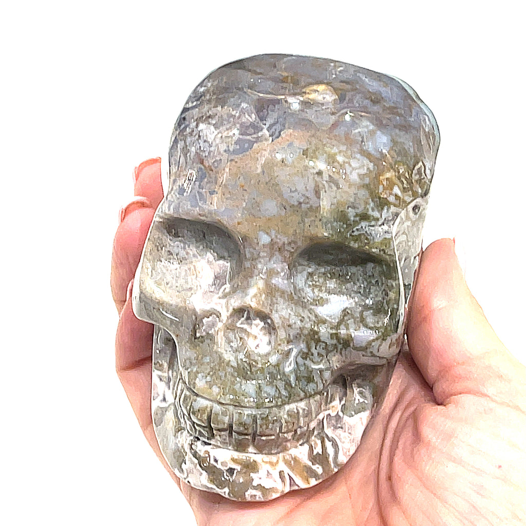 A carved gemstone skull, predominantly white in color with green accents, showcasing realistic facial features on a white background, front angle view in a models hand. 