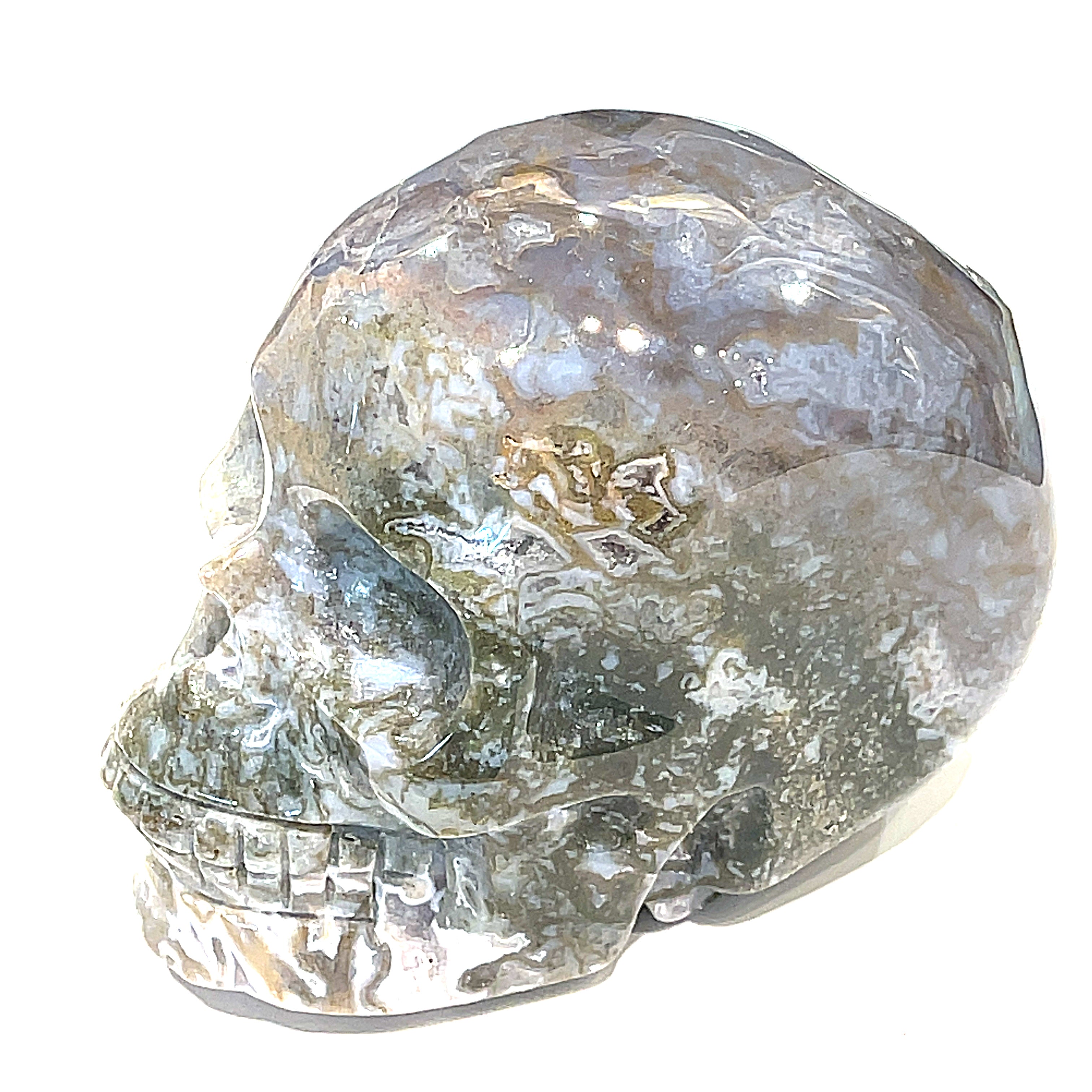 A carved gemstone skull, predominantly white in color with green accents, showcasing realistic facial features on a white background, right side view in a models hand. 