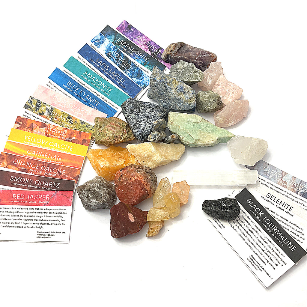 A collection of various raw gemstones is spread out, each with a small information card indicating the type of gemstone. The gemstones are colorful and displayed against a white background in a front overview.