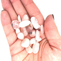A collection of xs tumbled Petalite gemstones with a pink hue, displayed  in a models palm on a white background, front overview.