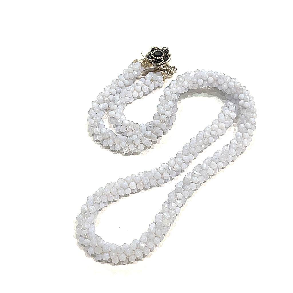 A crocheted necklace made with blue chalcedony gemstones, featuring a sterling silver flower box clasp and a black gemstone detail on a white background, long, front, close up overview.