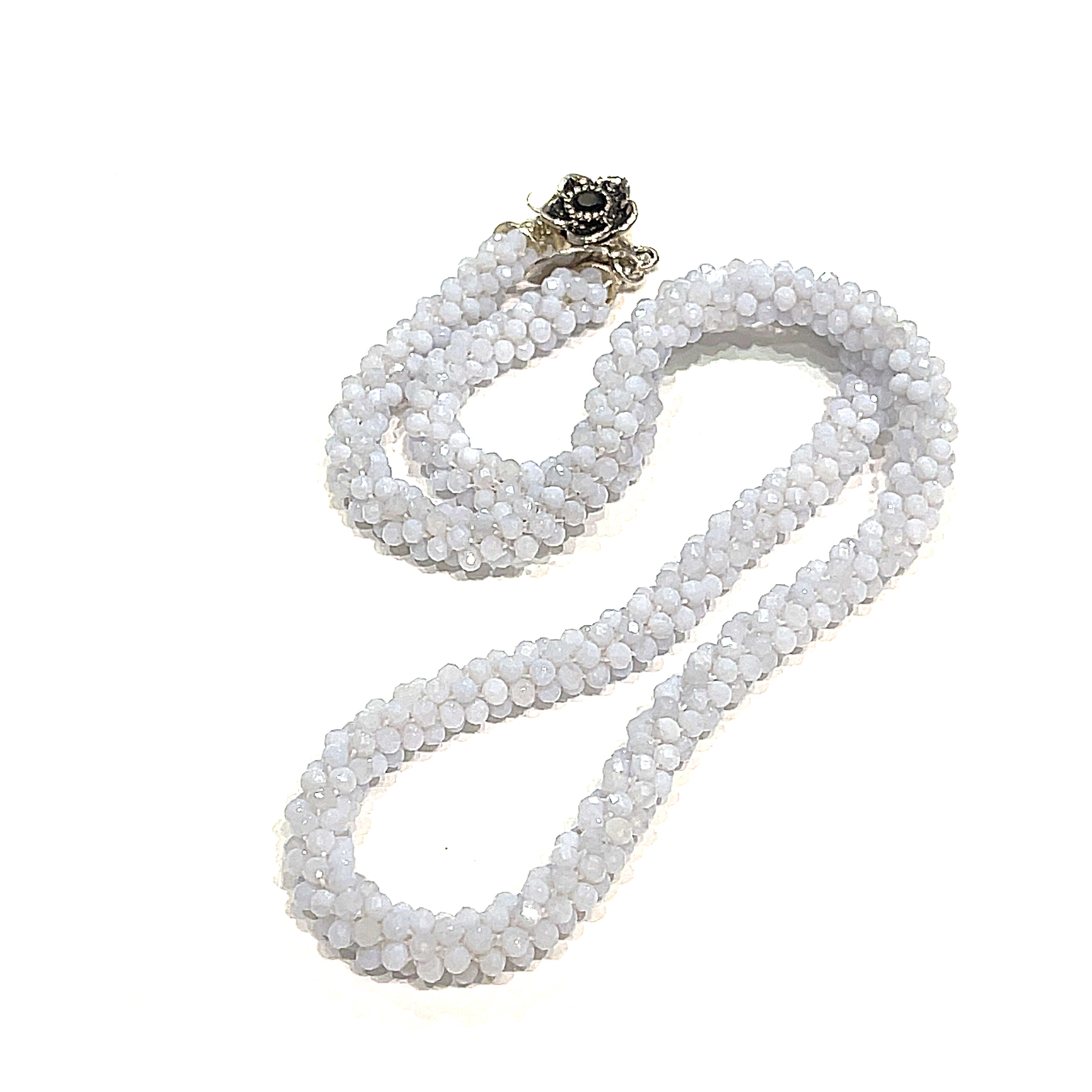 A crocheted necklace made with blue chalcedony gemstones, featuring a sterling silver flower box clasp and a black gemstone detail on a white background, long, front, close up overview.