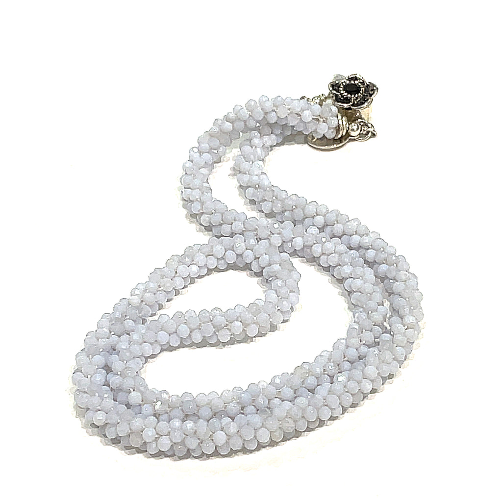 A crocheted necklace made with blue chalcedony gemstones, featuring a sterling silver flower box clasp and a black gemstone detail on a white background, long, front overview. 