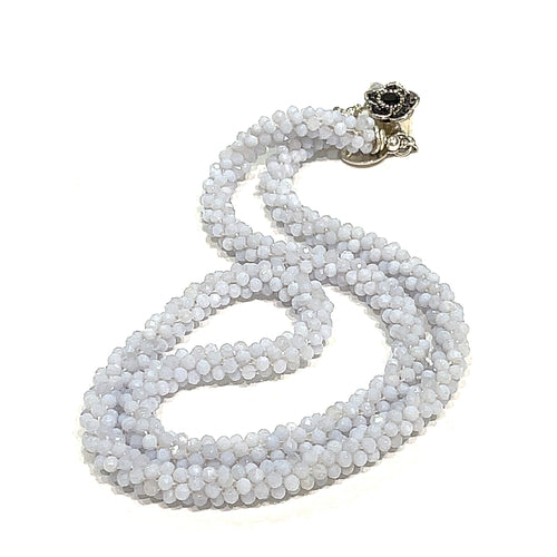 A crocheted necklace made with blue chalcedony gemstones, featuring a sterling silver flower box clasp and a black gemstone detail on a white background, long, front overview. 