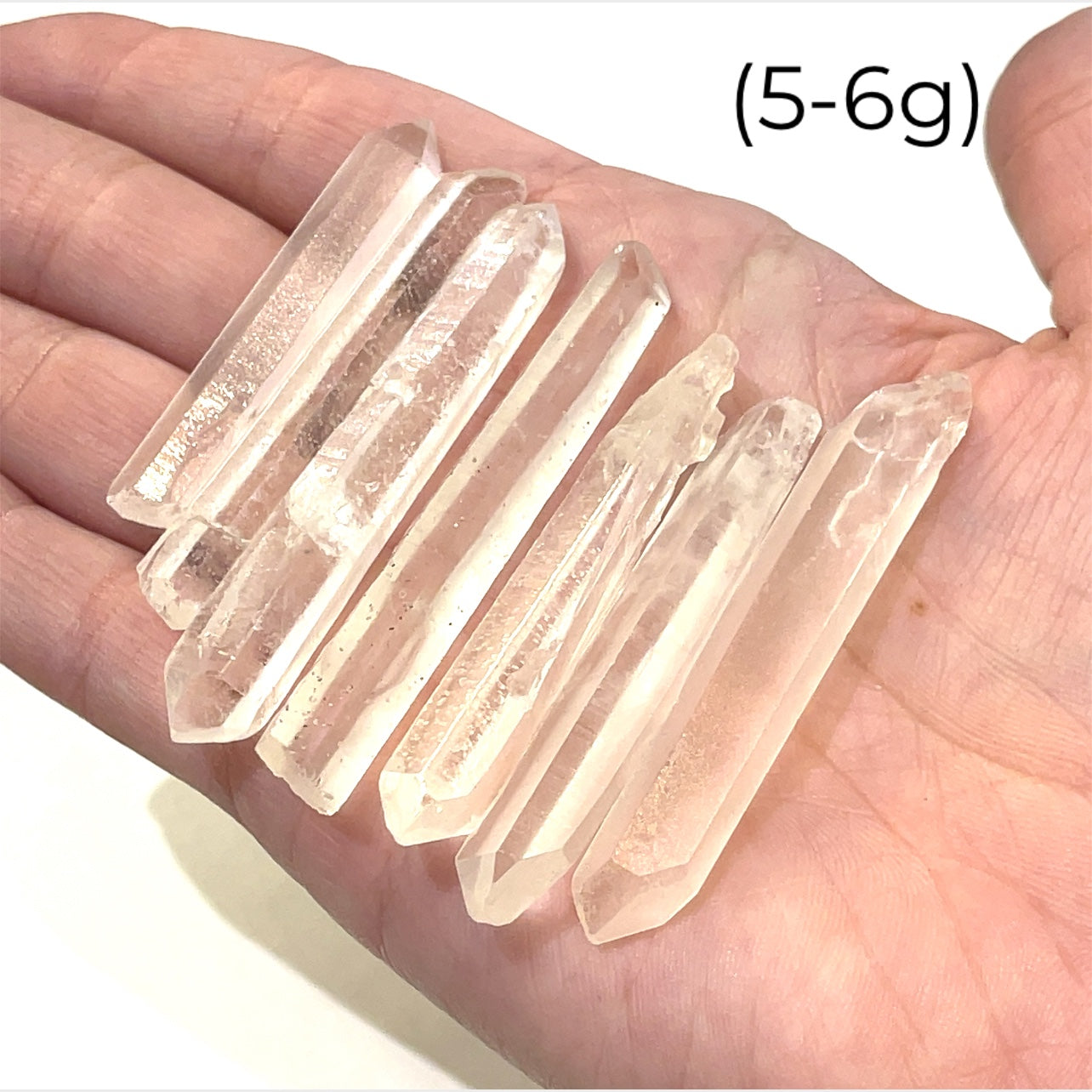 A hand holding a group of (5-6g) of clear quartz gemstone wands with a white background, front view.