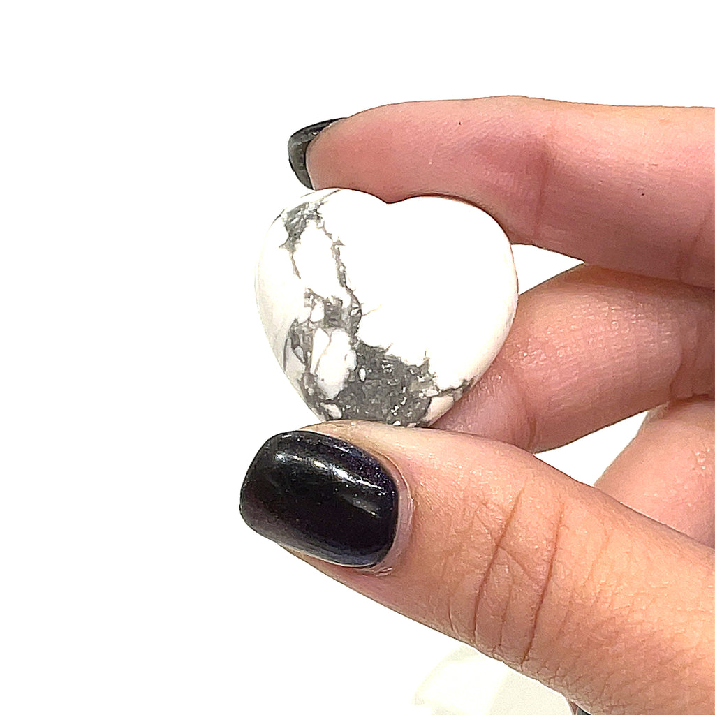 A hand holding a heart-shaped Howlite gemstone with a white and gray marbled pattern on a white background, front view. 