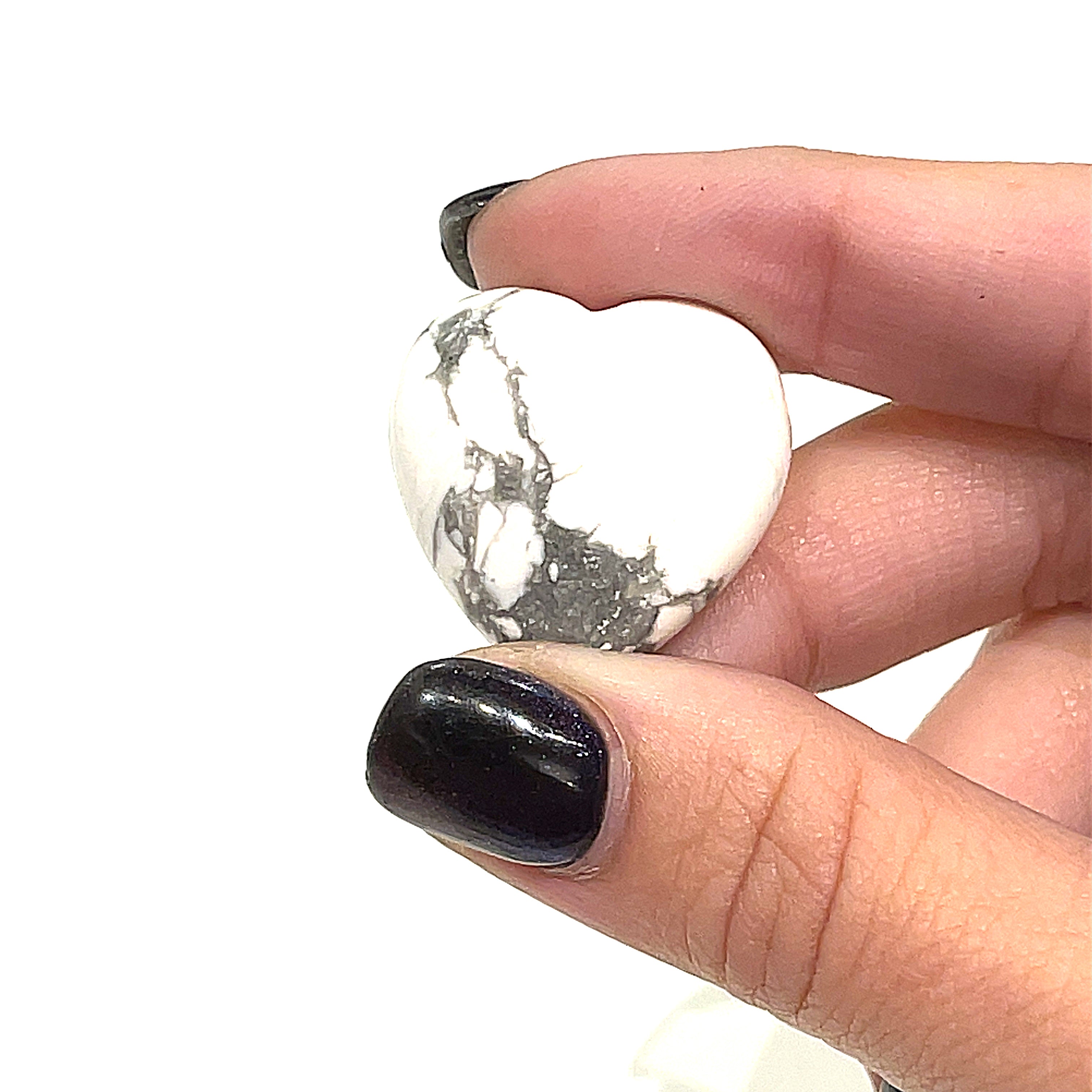 A hand holding a heart-shaped Howlite gemstone with a white and gray marbled pattern on a white background, front view. 