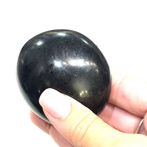 A hand holding a polished Black Tourmaline gemstone palm stone on a white background, side view. 