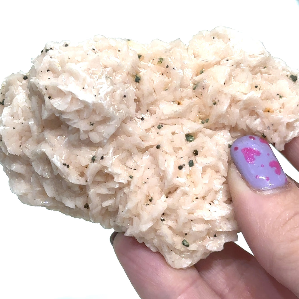 A hand holding a raw specimen of Pink Dolomite with Chalcopyrite, showing a rough, crystalline texture with specks of color on a white background, in a close-up view.
