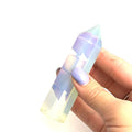 A hand holding a transparent multicolored opalite gemstone tower against a white background, front view. 