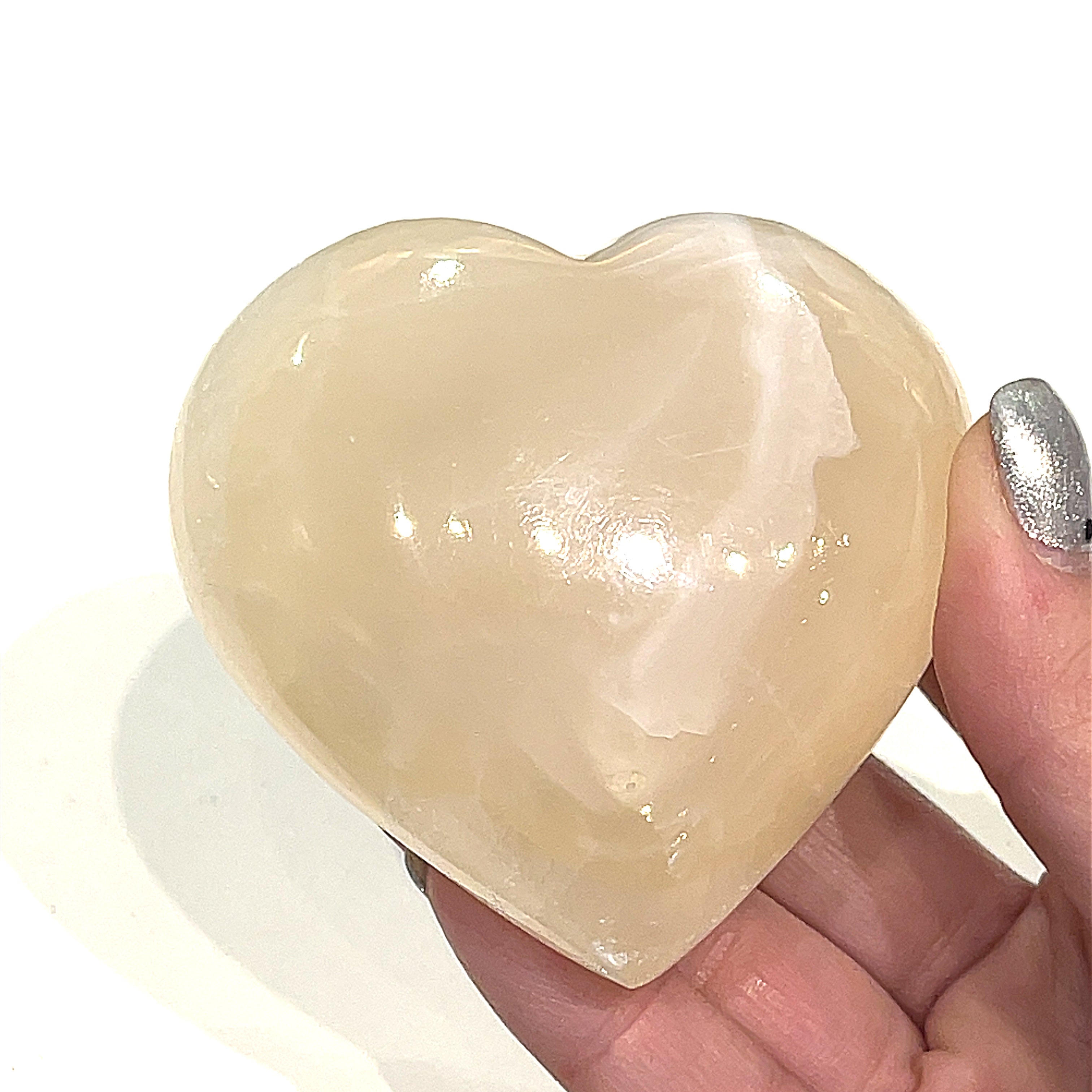 A hand holding an Lg pink Mangano Calcite gemstone heart on a white background, front view.