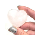 A hand holding an med pink Mangano Calcite gemstone heart on a white background, front view.