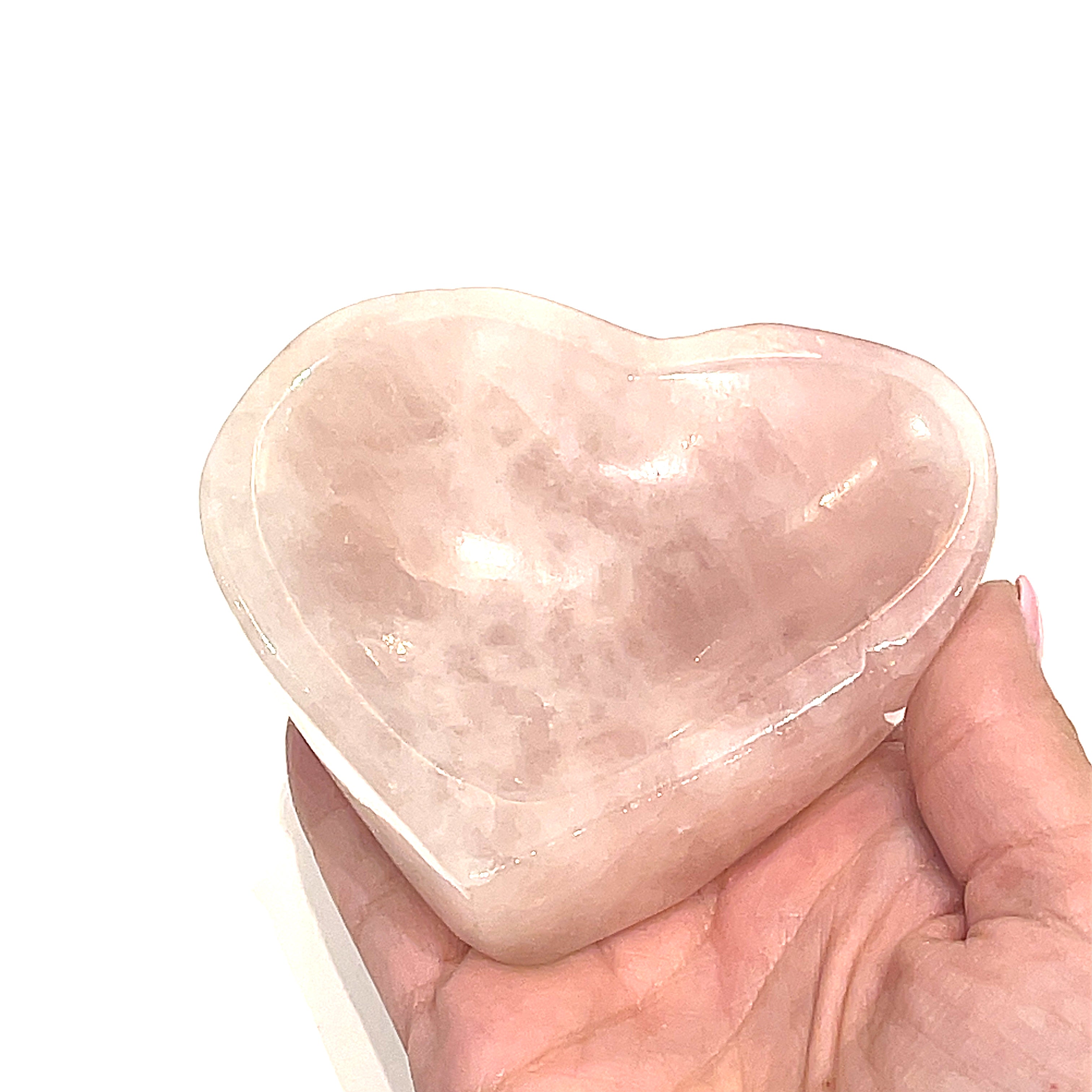 A heart-shaped Rose Quartz gemstone bowls, one pink, is placed in a models hand, front overview on a white background. 