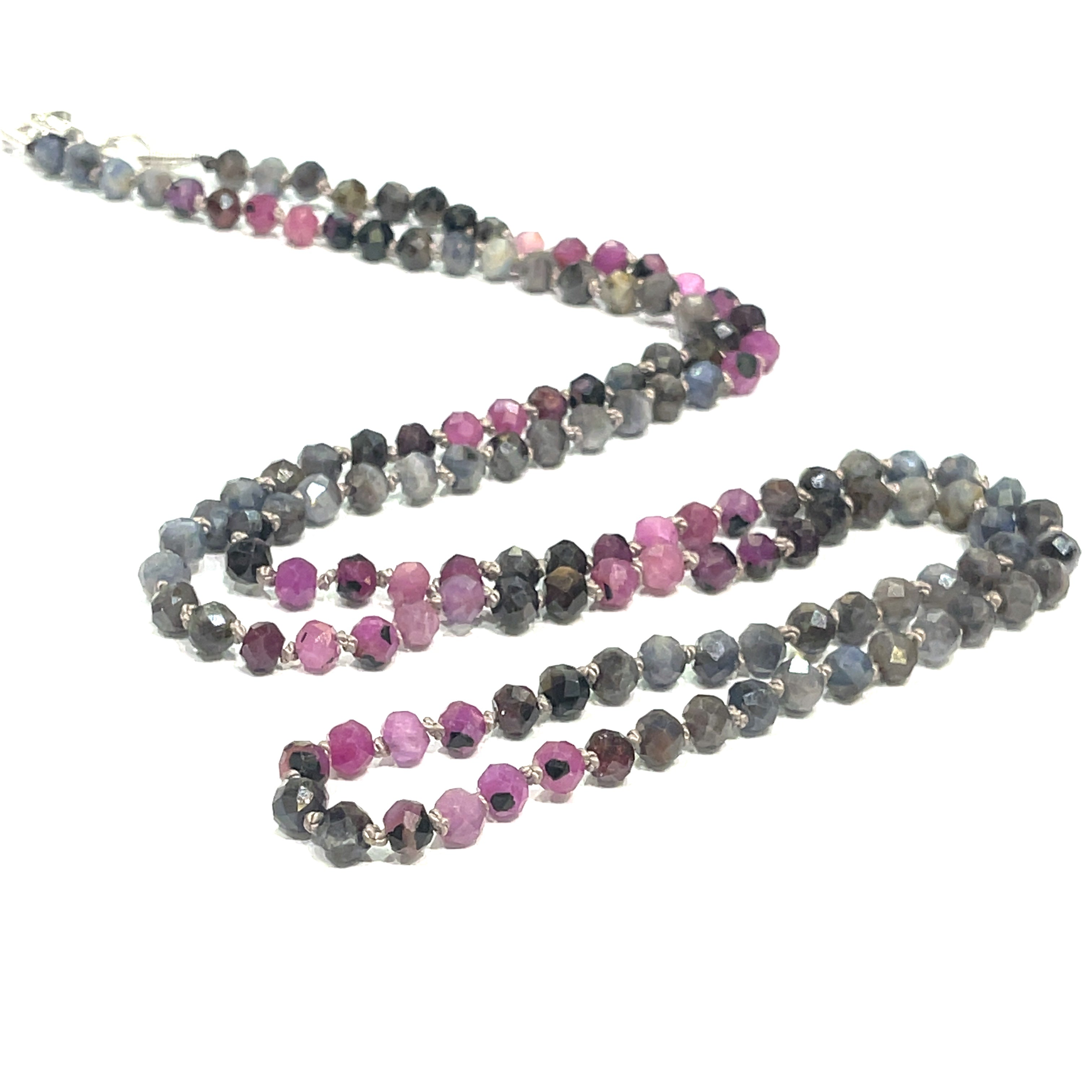 A knotted necklace featuring a heart lobster clasp, composed of faceted roundel rubies and sapphire gemstones on a white background, diagonal left  the center close up overview. 