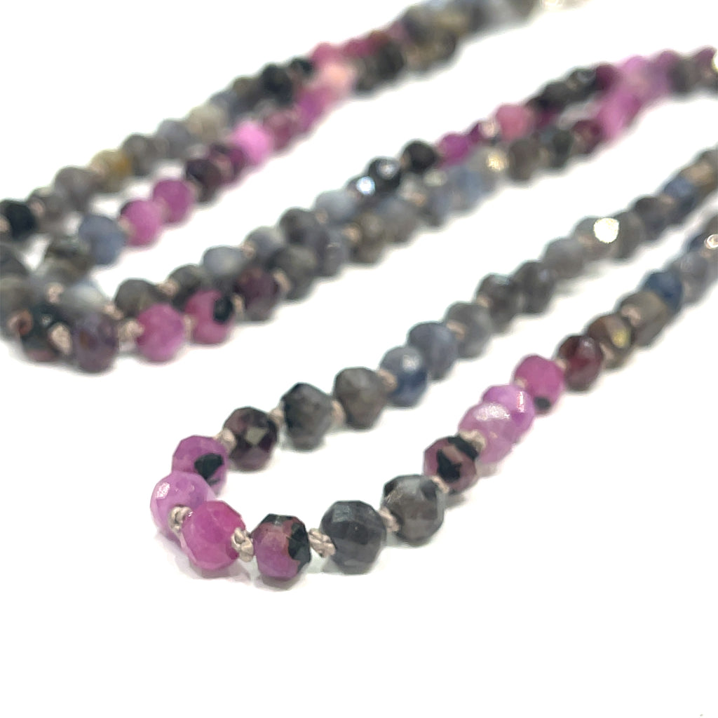 A knotted necklace featuring a heart lobster clasp, composed of faceted roundel rubies and sapphire gemstones on a white background, long center close up overview. 