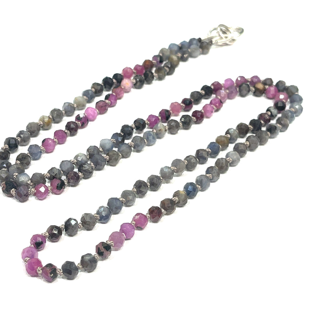 A knotted necklace featuring a heart lobster clasp, composed of faceted roundel rubies and sapphire gemstones on a white background, long center view. 