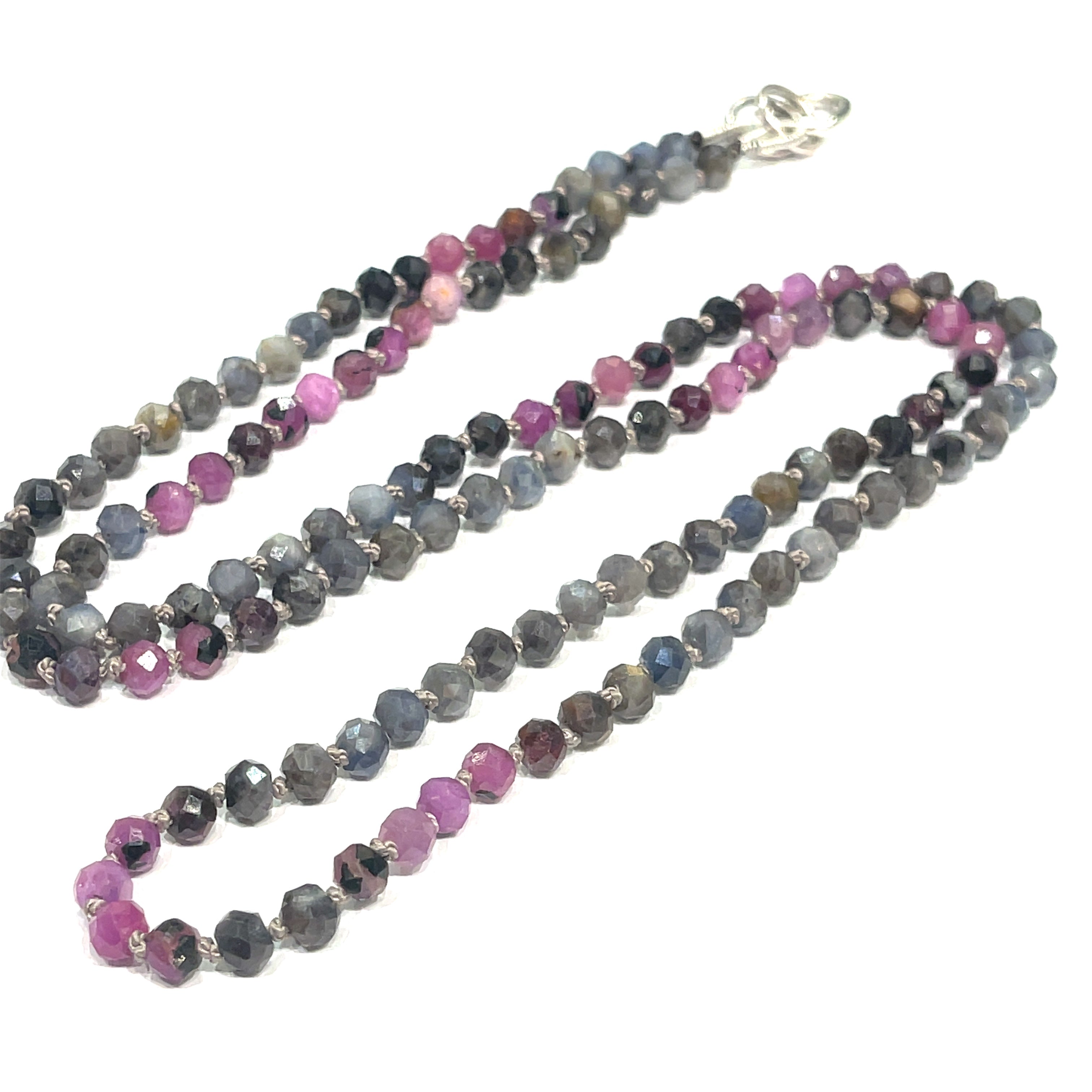 A knotted necklace featuring a heart lobster clasp, composed of faceted roundel rubies and sapphire gemstones on a white background, long center view. 