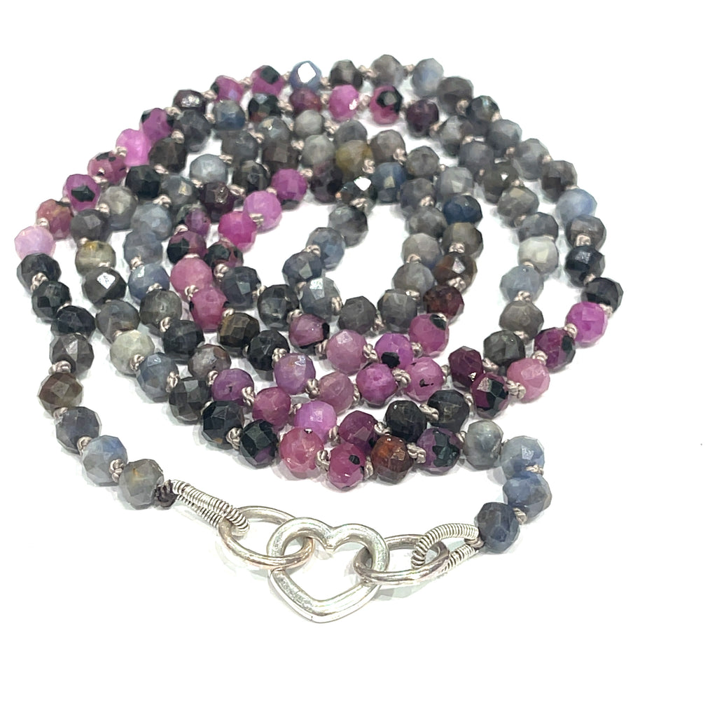 A knotted necklace featuring a heart lobster clasp, composed of faceted roundel rubies and sapphire gemstones on a white background, spiral center close up overview. 