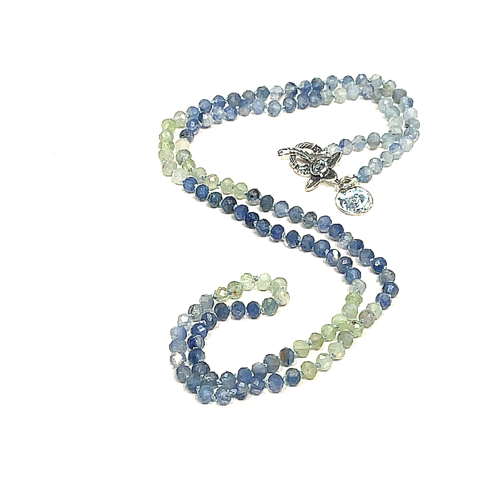 A knotted necklace featuring faceted roundel blue and green kyanite gemstone beads, with a sterling silver flower and a blue topaz gemstone toggle, on a white background, front-center overview. 
