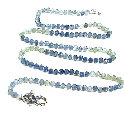 A knotted necklace featuring faceted roundel blue and green kyanite gemstone beads, with a sterling silver flower and a blue topaz gemstone toggle on a white background, front center, long overview. 