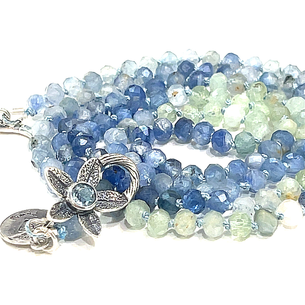 A knotted necklace featuring faceted roundel blue and green kyanite gemstone beads, with a sterling silver flower and a blue topaz gemstone toggle on a white background, front center overview. 